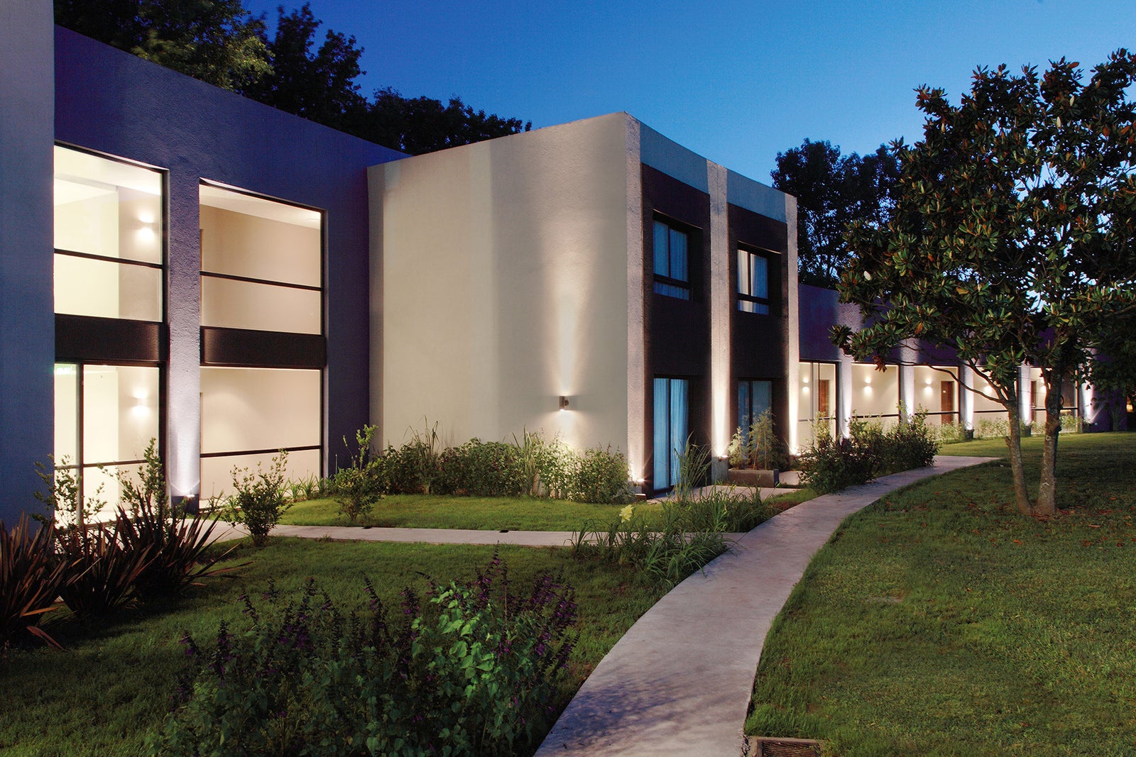 Modern illuminated building at dusk with a curved walkway, landscaping, and trees.