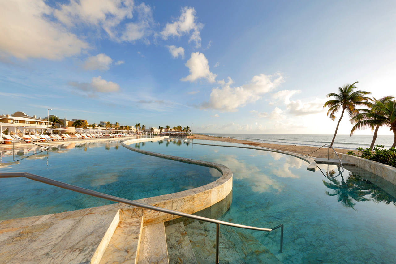 Beachfront resort with interconnected turquoise pools, palm trees, lounge chairs, and ocean views under a partly cloudy sky.