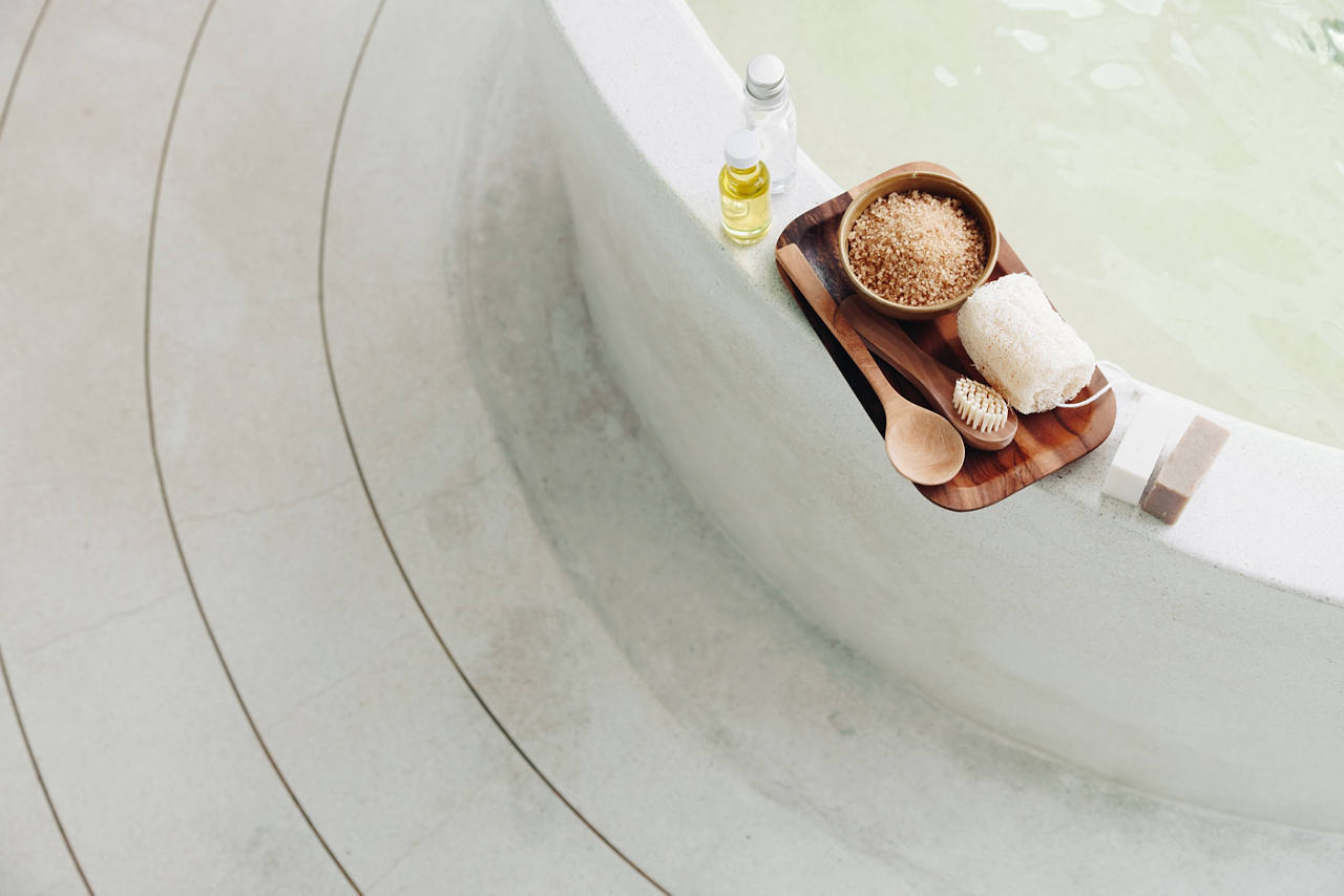 Spa bath with a wooden tray of bath salts, towel, toiletries, brush, spoon, and soap resting on the rim of a curved white pool.