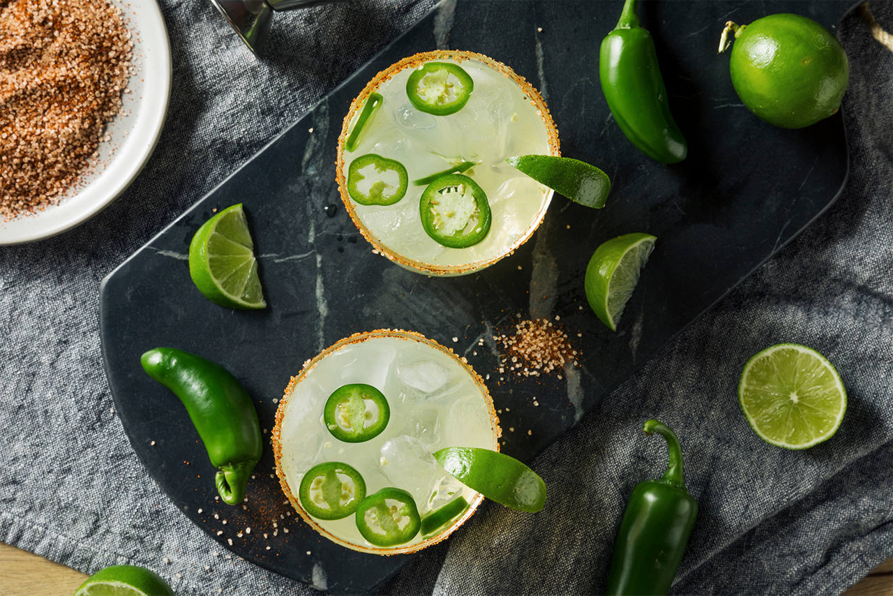 Overhead arrangement of two lime- and jalapeño-garnished drinks with citrus, peppers, and seasoning on a dark slate surface.