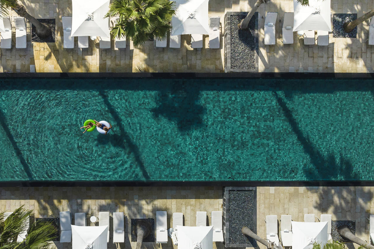 Overhead view of a turquoise swimming pool with a person floating near the left side and long shadows cast across the water.
