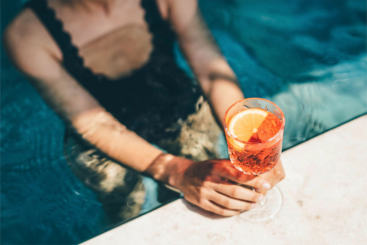 Person in a pool holding a glass of orange-red iced drink garnished with an orange wedge.