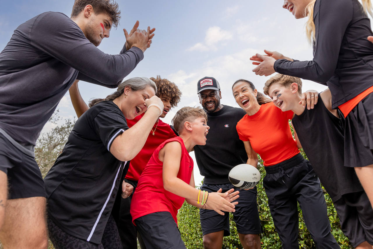 A group of diverse adults and children gather outdoors in a loose circle smiling and motivated around a rugby-style ball.