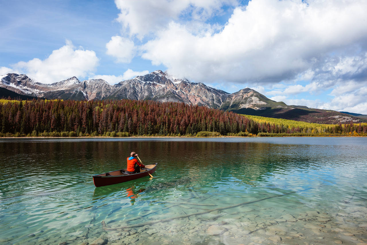 A kayak with a rider on a crystal clear lake, overlooking a mountain landscape with white clouds against a blue sky.