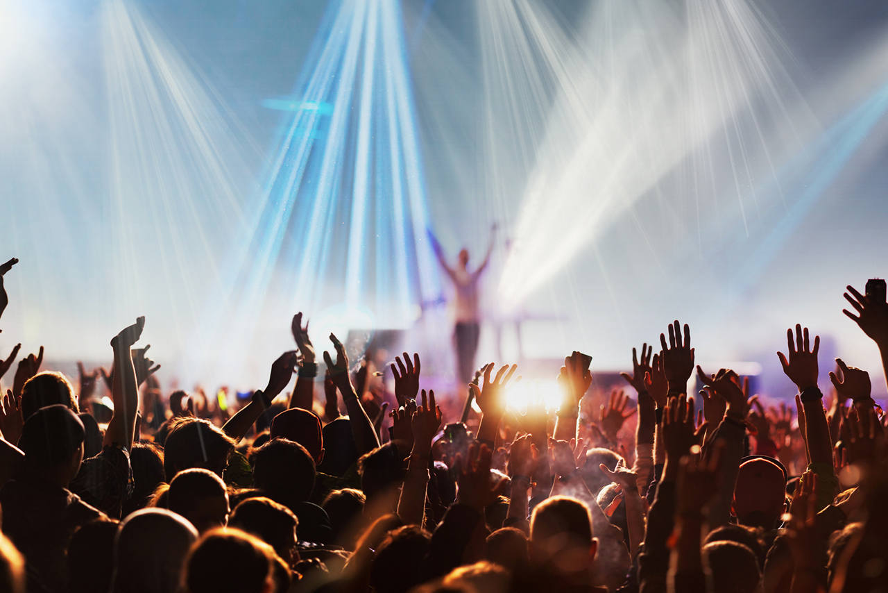 View of a crowded concert with a stage lit by beams of light with a song artist standing in the middle of it.