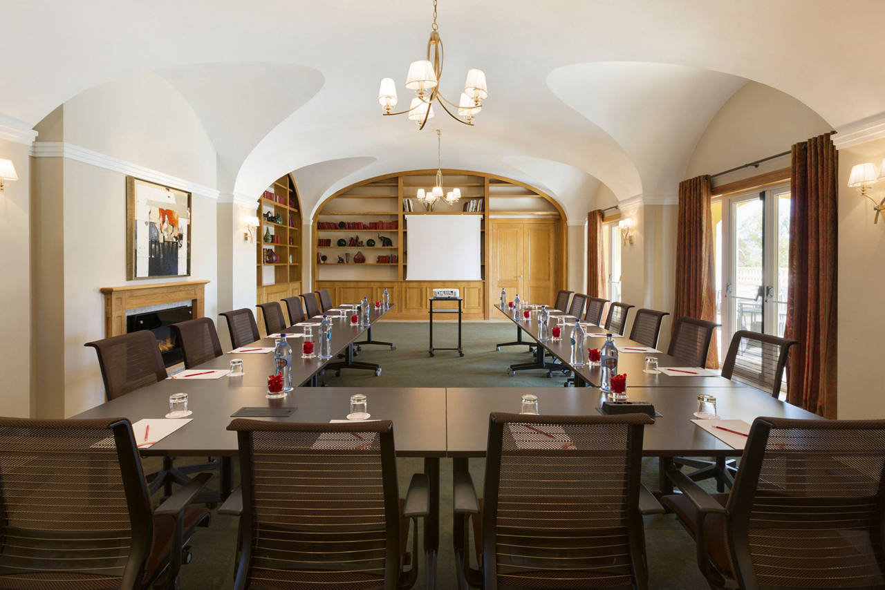 An elegant conference room with a fireplace on one side and french, curtained double doors in the other.  In the middle is a U-shaped table lined with ergonomic chairs, each appointed with a pad, pen, and a drinking glass, all facing a screen with a projector. 
