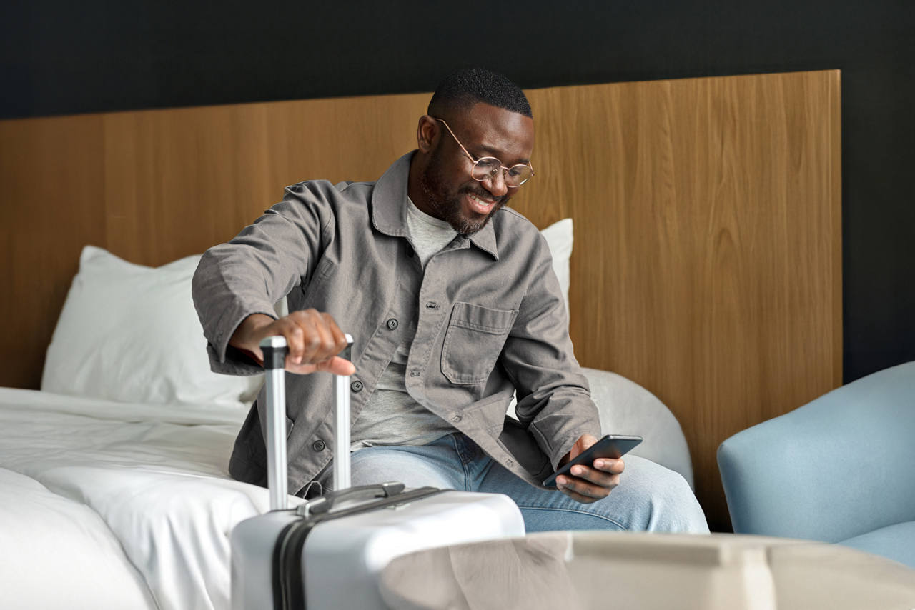 A man sitting on his hotel bed ready to leave with his packed luggage, digitally checking out on his phone.